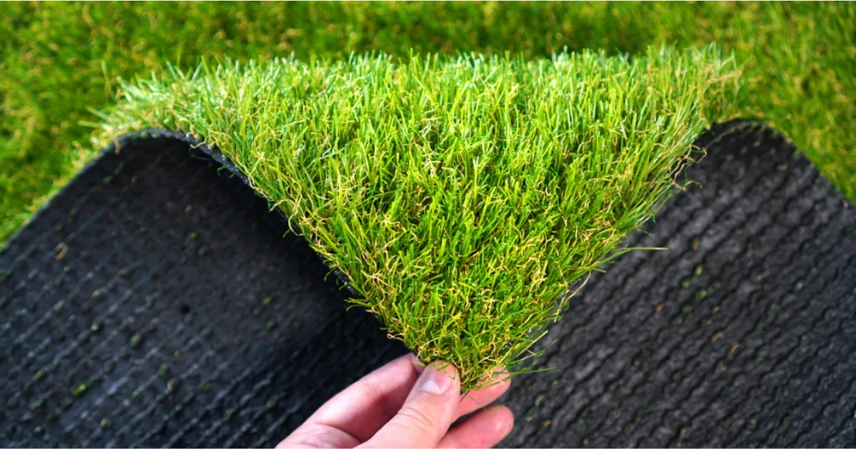 A hand lifting the edge of a piece of artificial grass to reveal the black backing material beneath.